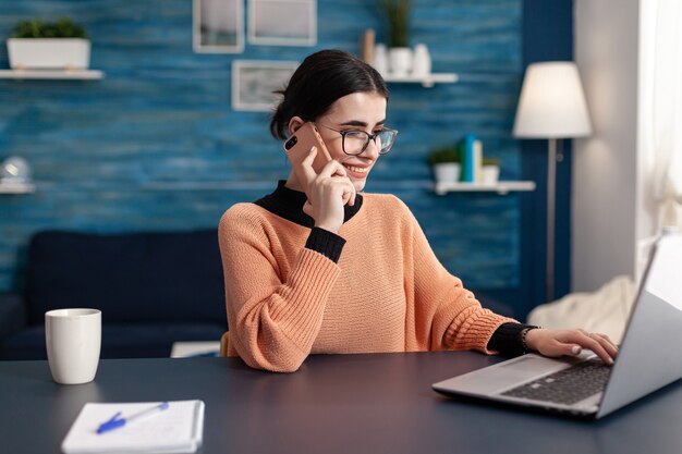 Adolescente, estudando no projeto de comunicação para o seminário universitário, usando o computador portátil. Aluno falando ao telefone com o colega enquanto está sentado à mesa da escrivaninha na sala de estar