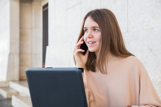 Adolescente feminino positivo usando o laptop e telefone na parede do edifício