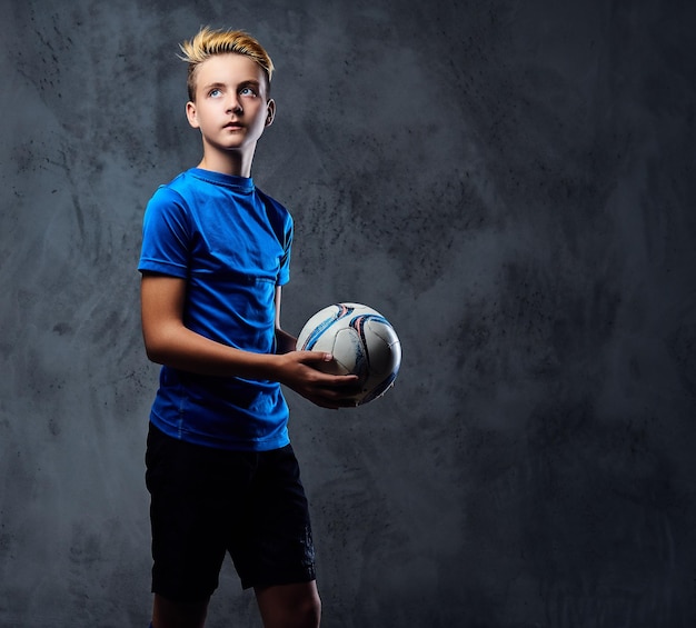 Foto grátis adolescente loiro, jogador de futebol vestido com um uniforme azul segura uma bola.