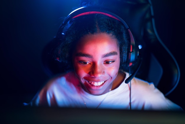 Foto grátis adolescente negra sorridente em fone de ouvido jogando videogame em clube de videogame com iluminação azul e vermelha