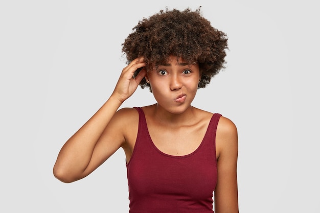 Adolescente negro, hesitante e sem noção, tem uma expressão facial incerta, curva os lábios, mantém a mão na cabeça, pondera sobre algo