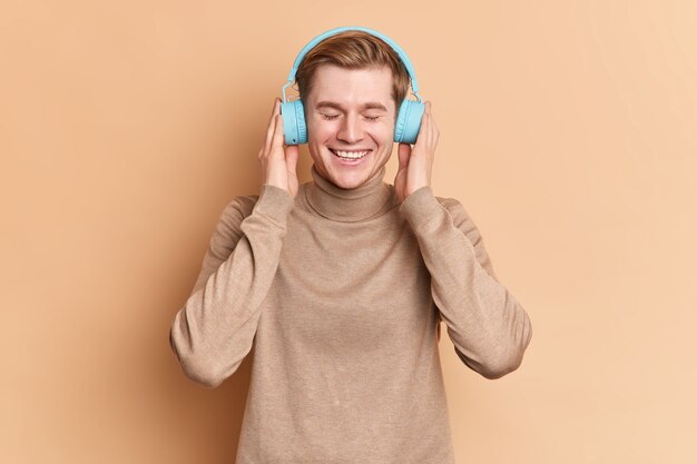 Adolescente satisfeito relaxa com os olhos fechados ouve a música favorita com fones de ouvido azuis sem fio usa app de música sorri com alegria usa poses casuais