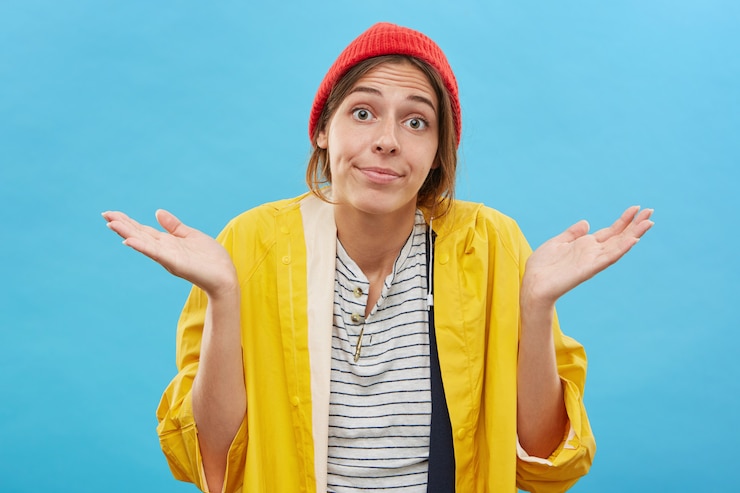Foto grátis adolescente sem noção usando chapéu vermelho estiloso e capa de chuva amarela olhando com expressão de olhos arregalados no rosto, encolhendo os ombros em ignorância ou indiferença, dizendo: as coisas acontecem
