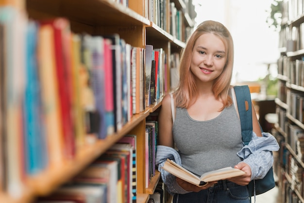 Adolescente sorridente com livro encostado na estante