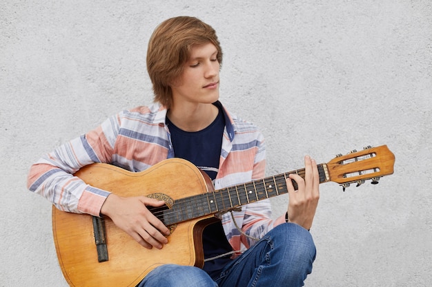Adolescente talentoso com penteado moderno, segurando o violão tocando suas músicas favoritas enquanto está sentado contra a parede de concreto cinza