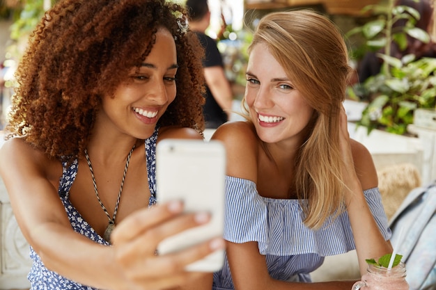 Foto grátis adoráveis mulheres de diferentes nacionalidades posam para selfie e se divertem juntas, têm expressões positivas, usam tecnologias modernas para se entreter, postam fotos para baixar nas redes