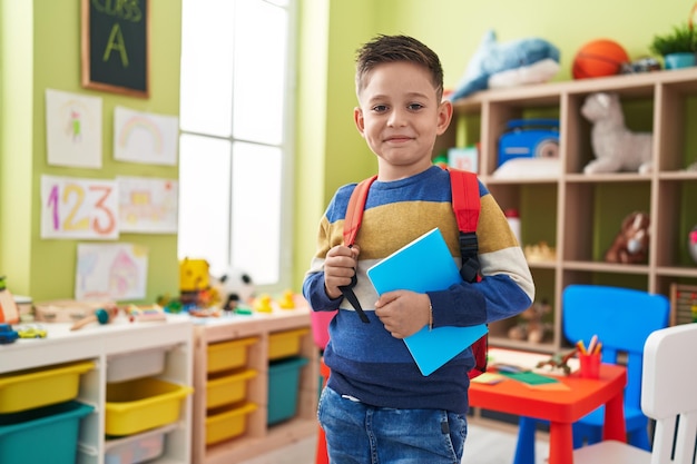 Adorável estudante hispânico usando mochila segurando livro no jardim de infância
