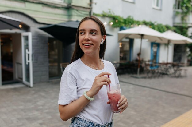 Adorável garota adorável em camiseta branca com smoothie está olhando para longe e sorrindo na rua brilhante de verão na cidade Garota feliz está andando na rua em bom dia