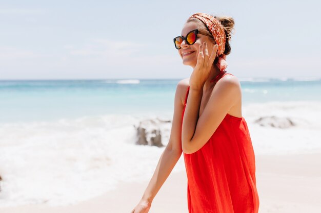 Adorável garota caucasiana, passando o verão em um lugar exótico perto do mar. Foto ao ar livre de uma senhora sorridente graciosa de óculos escuros, posando na praia