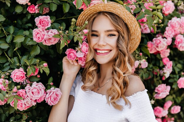 Adorável garota com cabelo loiro encaracolado, posando no jardim. Retrato de mulher feliz caucasiana segurando flor rosa.