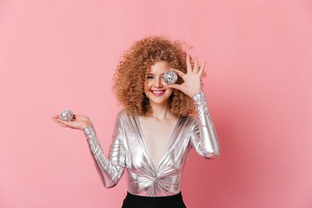 Adorável garota com cachos loiros, vestida com blusa prata, está sorrindo e posando com bolas de discoteca no espaço rosa.