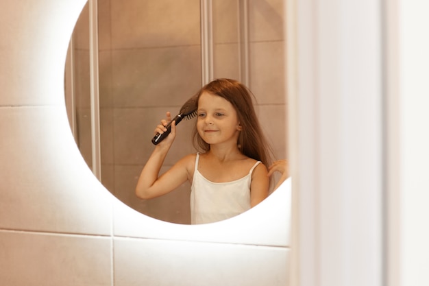 Foto grátis adorável garota de cabelos escuros penteando o cabelo no banheiro em frente ao espelho, olhando seu reflexo, fazendo procedimentos de beleza matinais, expressando felicidade.