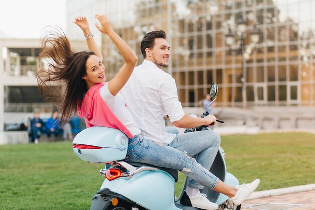 Adorável garota de camisa rosa, olhando por cima do ombro enquanto andava de scooter com o namorado. Foto ao ar livre da parte de trás do casal dirigindo pela cidade durante as férias.