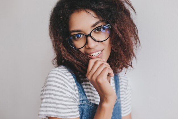 Adorável garota negra com penteado da moda posando divertidamente na parede de luz, olhando através de óculos elegantes