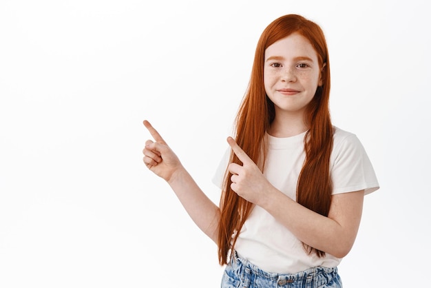 Adorável garota ruiva com sardas, criança ruiva, apontando os dedos no canto superior esquerdo, sorria tímido em pé sobre fundo branco em camiseta casual
