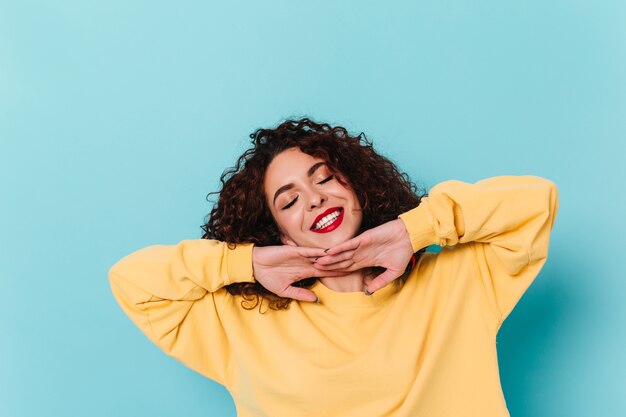 Adorável garota vestida com um suéter amarelo toca seu rosto e sorri com os olhos fechados contra o espaço azul.