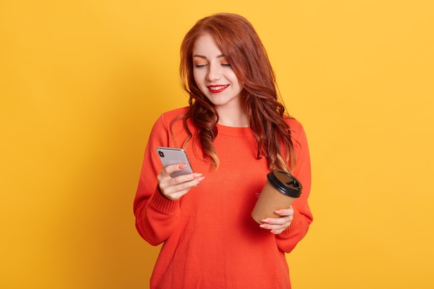 Adorável garota vestindo suéter laranja posando isolada, segurando café para levar e o celular