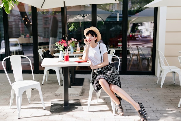 Adorável jovem com chapéu de verão descansando em um café ao ar livre com a mão apoiada no rosto e um amigo esperando