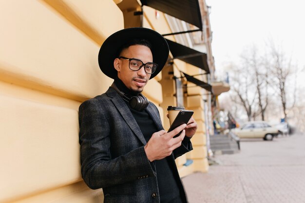 Adorável jovem com pele morena em pé perto de um prédio antigo com uma xícara de café. Modelo masculino africano na moda em traje casual, segurando o telefone enquanto posava na rua.