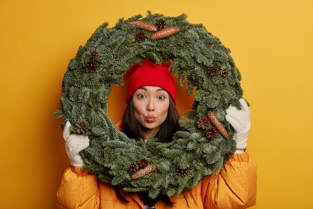 Adorável jovem coreana mantém os lábios arredondados, olha através da coroa de abetos verdes, usa casaco amarelo e luvas brancas, decora a casa antes do Natal, posa em ambientes fechados.