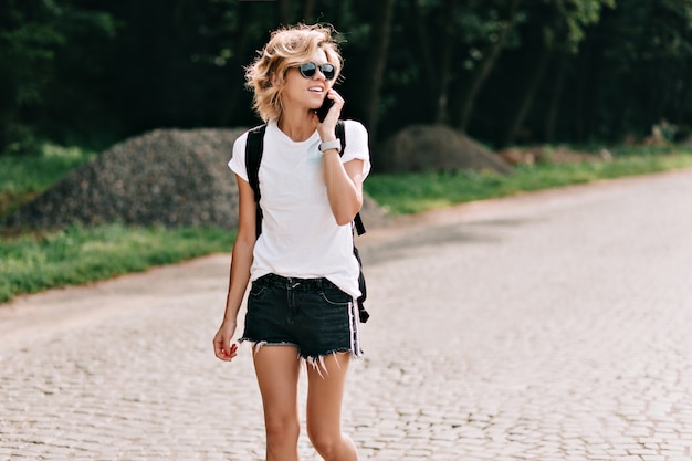 Adorável jovem encantadora com penteado curto, andando na estrada com uma mochila e falando ao telefone sobre as montanhas. Humor de viagens, férias, viagem. desejo de viajar e conceito de viagens.