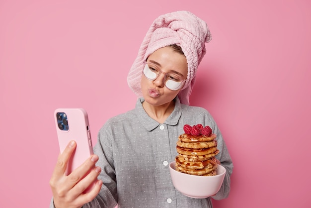 Adorável jovem faz selfie inclina a cabeça mantém os lábios arredondados poses na câmera do smartphone posa com panquecas preparadas com calda e framboesas usa toalha de pijama enrolada na cabeça Despertar feliz