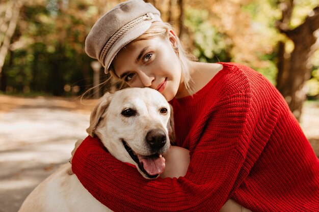 Adorável jovem num belo pulôver vermelho na moda abraçando labrador na floresta. Loira bonita com chapéu leve com seu cachorro sentado no parque.