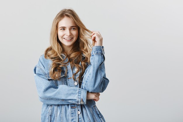 Adorável loira sorrindo feliz em uma jaqueta jeans, em pé sobre uma parede cinza