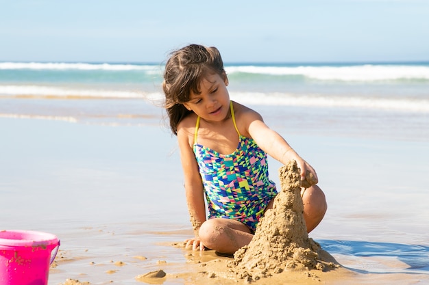 Adorável menina construindo castelo de areia na praia, sentada na areia molhada, aproveitando as férias à beira-mar