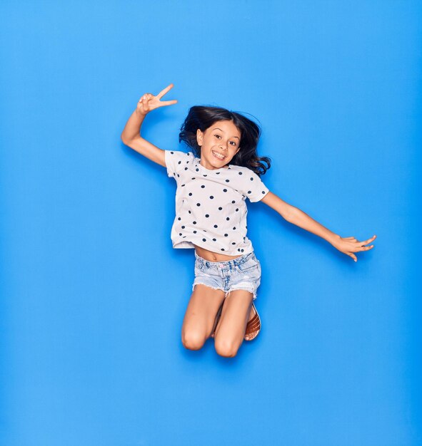 Adorável menina hispânica vestindo roupas casuais sorrindo feliz pulando com sorriso no rosto fazendo sinal de vitória sobre fundo azul isolado