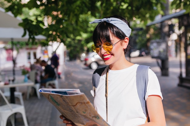 Adorável menina morena com sorriso hollywood olhando para o mapa da cidade em busca de um destino parada no meio da rua