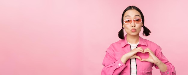 Adorável modelo feminino coreano em óculos de sol da moda mostra sinal de cuidado cardíaco eu te amo gesto fica sobre fundo rosa