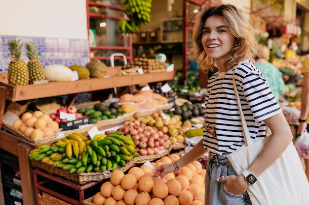 Adorável mulher adorável com cabelos claros ondulados vestindo camiseta listrada andando no mercado de frutas e escolhendo frutas e legumes Garota sorridente feliz está comprando comida ecológica