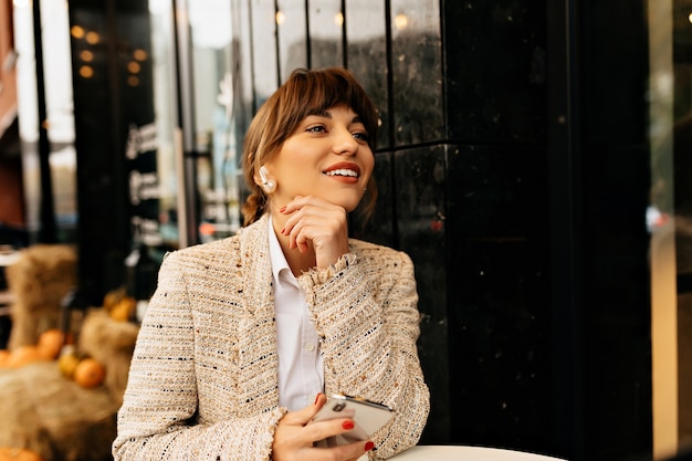Adorável mulher adorável em roupas da moda sentada em um café ao ar livre, usando o smartphone e esperando por amigos no fundo das luzes da cidade