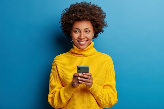 Adorável mulher adulta de pele escura vestida com um macacão amarelo usando um telefone celular com uma expressão feliz