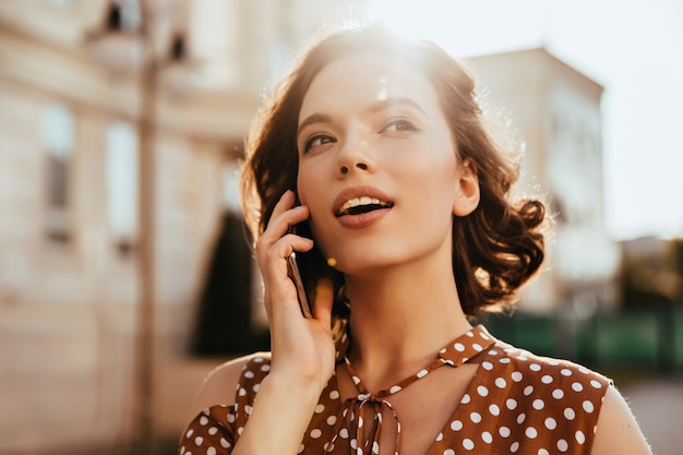 Adorável mulher branca ligando para alguém enquanto caminhava pela cidade. Tiro ao ar livre da sonhadora garota caucasiana em pé na rua com o celular.