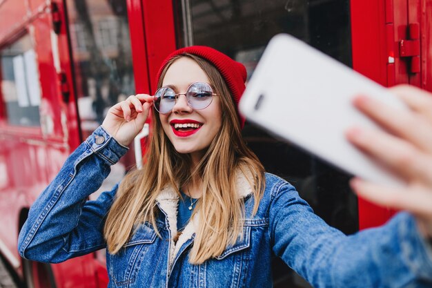 Adorável mulher caucasiana brincando de tocar seus óculos enquanto faz selfie perto do ônibus.