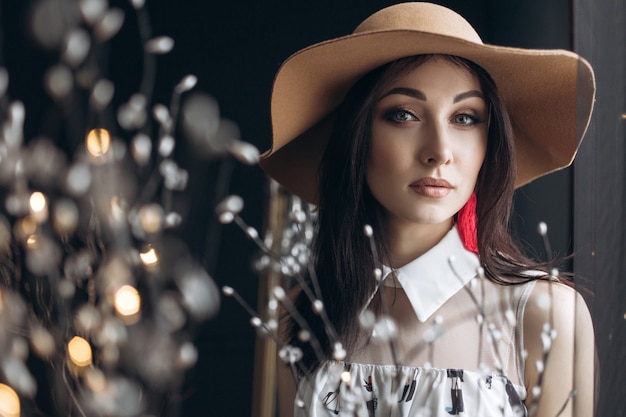 Adorável mulher de chapéu bege e com olhar de moda poses no estúdio escuro