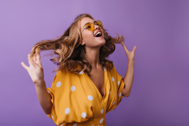 Adorável mulher europeia brincando na violeta. Menina bronzeada na moda brincando com seu cabelo loiro ondulado.