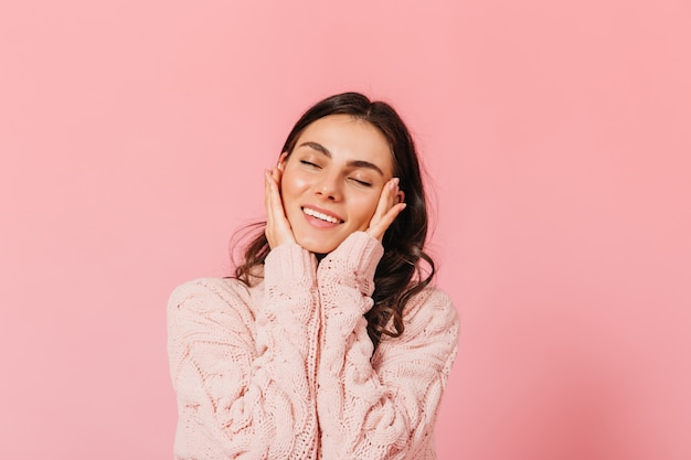Adorável mulher morena sorri ternamente com os olhos fechados. Senhora de suéter quente posando no estúdio rosa.