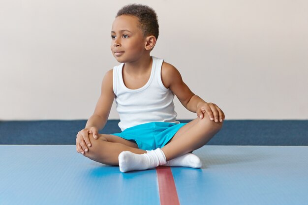 Adorável pequeno desportista de aparência africana sentado no tapete com as pernas cruzadas e relaxantes após um treino intensivo.