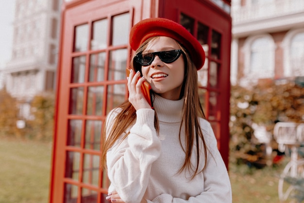 Adorável senhora com cabelo comprido, usando boné vermelho e óculos pretos, perto de uma cabine telefônica vermelha