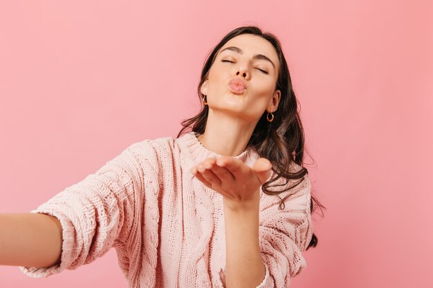 Adorável senhora de cabelos escuros na camisola de malha manda beijo e faz selfie no fundo rosa.