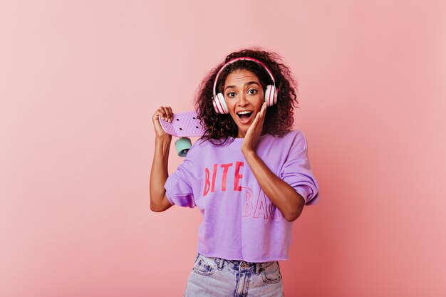 Adorável senhora de olhos escuros em fones de ouvido, posando com um sorriso surpreso. Linda garota africana com skate expressando espanto em rosa.