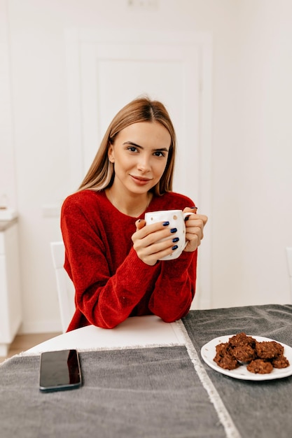 Adorável senhora elegante com sorriso feliz vestida de pulôver vermelho está segurando uma xícara de café e descansando em casa