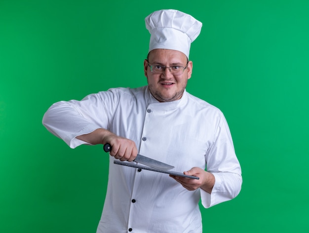 Adulto alegre cozinheiro masculino usando uniforme de chef e óculos olhando para frente tocando a tábua de corte com uma faca isolada na parede verde