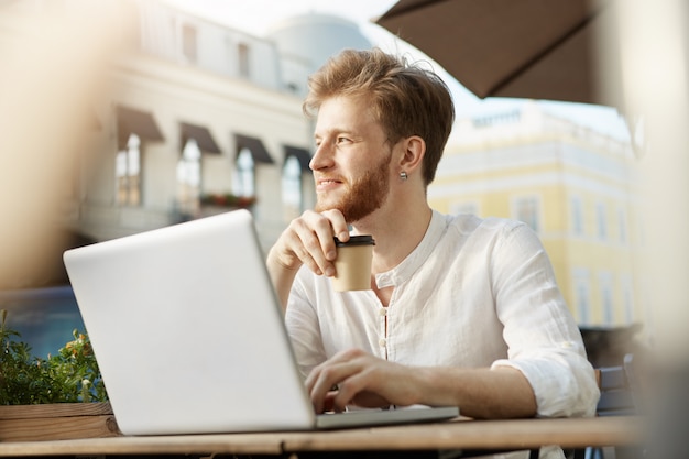 Adulto gengibre homem bonito com o computador portátil, sentado no terraço de um restaurante ou café