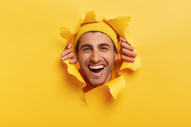 Adulto positivo do sexo masculino com barba por fazer parece feliz através do papel amarelo, mostra apenas o rosto