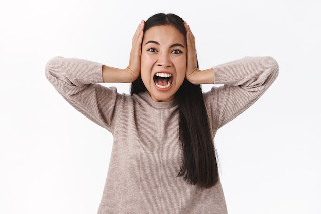 Aflito e farto, deprimido, alarmado, mulher furiosa, perdendo o controle, tendo colapso mental, gritando agarre a cabeça e olhe para a câmera com dor e raiva, grite furioso, fique de pé na parede branca