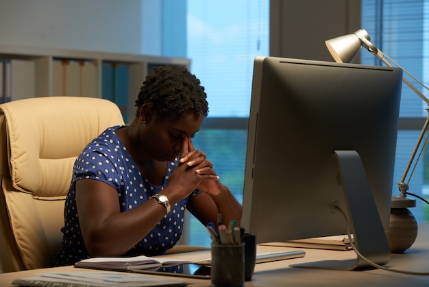 Afro-americana mulher sentada na frente do computador no escritório e inclinando a cabeça nas mãos entrelaçadas